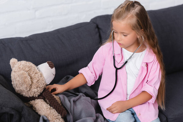 Child holding stethoscope near soft toy while sitting on sofa 