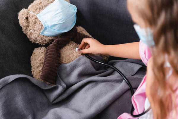 Selective focus of child holding stethoscope near teddy bear in medical mask 