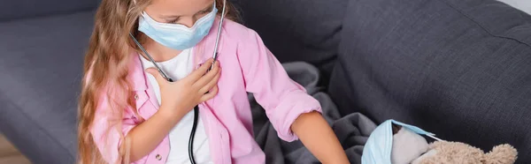 横の画像の子供で医療用マスクでStethoscope座って近くテディベアオンソファ — ストック写真