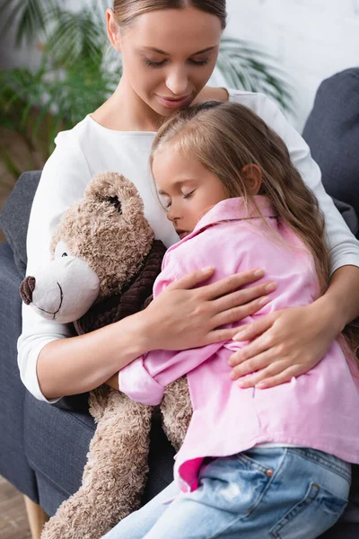 Selective focus of woman embracing sick child with teddy bear on couch