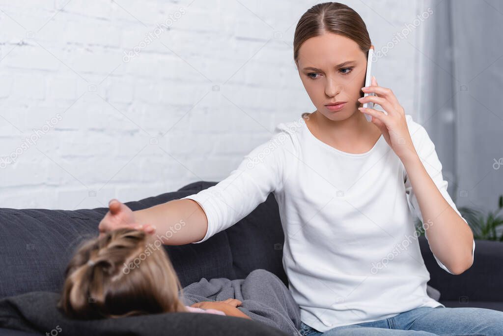 Selective focus of young woman talking on smartphone and touching forehead of ill daughter on sofa 