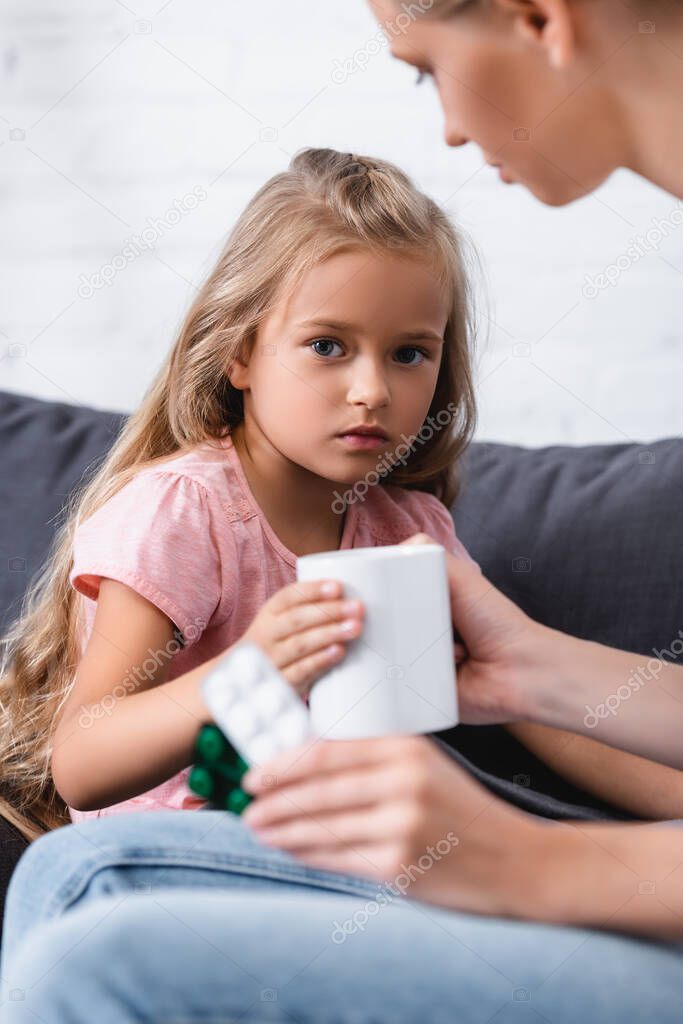 Selective focus of child looking at camera near mother with mug and pills at home 