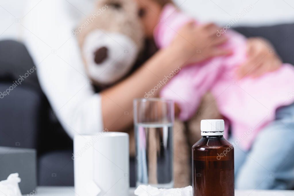 Selective focus of bottle of syrup, cup and glass of water near mother embracing kid 