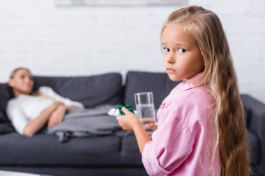 Selective focus of girl looking at camera while holding pills an glass of water near sick woman at home  clipart