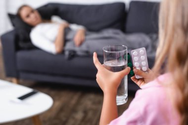Selective focus of child holding blisters with pills and glass of water near sick mother at home  clipart