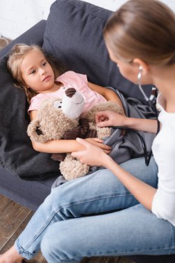Steteskoplu seçici bir kadın kanepesinde oyuncak ayıyla kızının yanında oturuyor. 