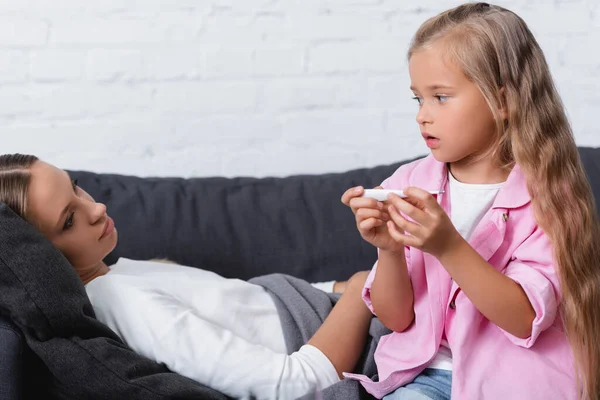 Ung Kvinna Liggande Soffan Och Tittar Barn Med Termometer — Stockfoto