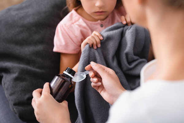 Cropped view of young woman pouring syrup near sick child holding blanket at home 