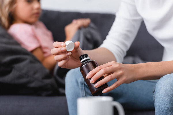Selective focus of woman holding syrup while sitting near sick daughter on couch 