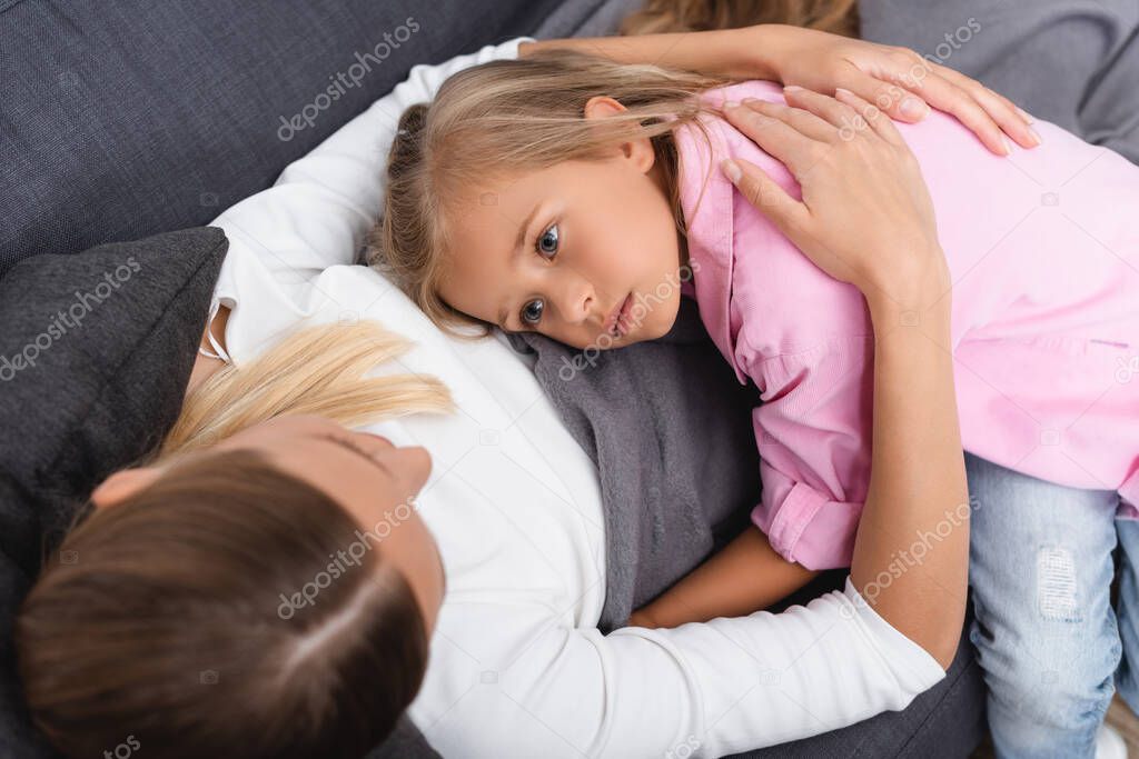 Overhead view of ill woman embracing daughter while lying on couch 