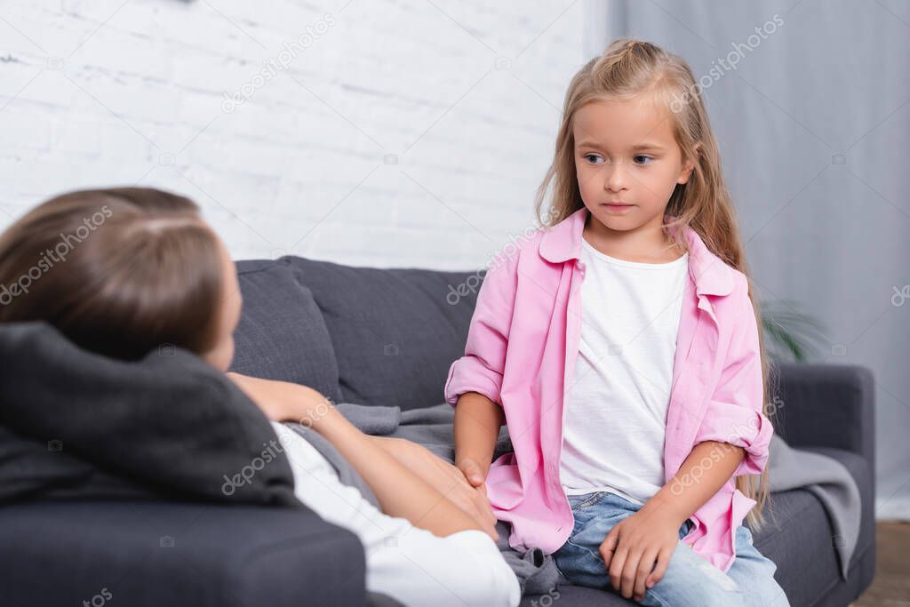 Selective focus of child holding hand of ill mother on couch in living room 