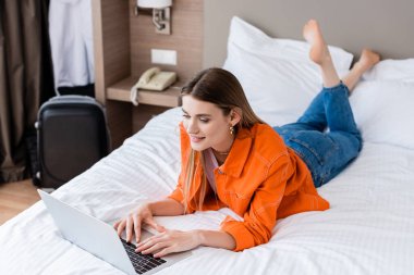 Yatakta yatıp otel odasında dizüstü bilgisayar kullanan çıplak ayaklı bir serbest yazar.