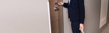 panoramic crop of businesswoman in suit holding room card while unlocking door in hotel  clipart