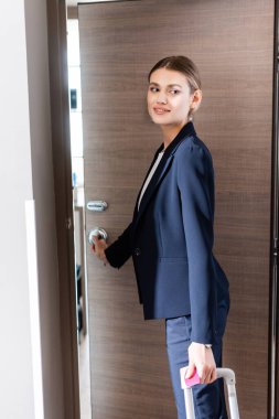 joyful businesswoman in suit opening door in hotel  clipart