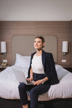 Takım elbiseli rüya gibi bir kadın otel odasında dizüstü bilgisayarla yatakta oturuyor. 