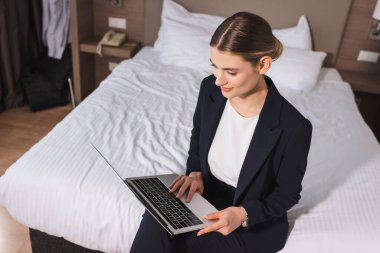 Yatakta oturan ve otel odasında dizüstü bilgisayar kullanan takım elbiseli kadının yüksek açılı görüntüsü. 