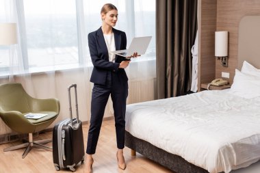 Otel odasındaki seyahat çantasının yanında duran ve laptop kullanan bir iş kadını. 