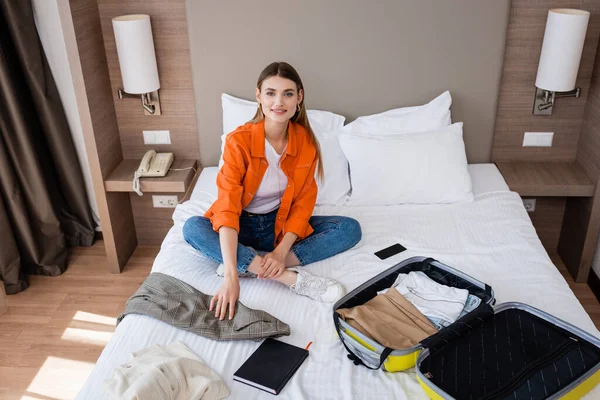 旅馆里的年轻女子坐在床上 身边有衣服 黄色行李 智能手机和笔记本 — 图库照片