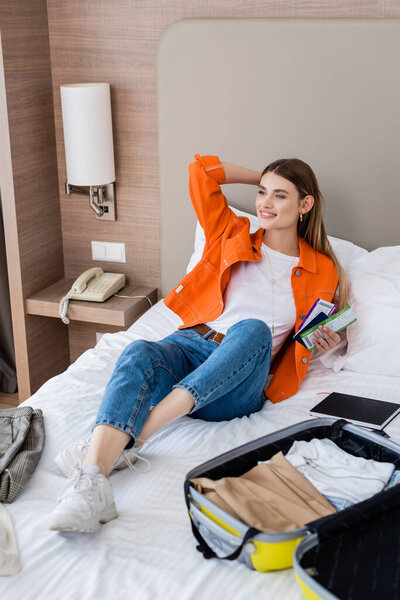 woman holding passport, boarding pass and air ticket near baggage and notebook on bed in hotel 
