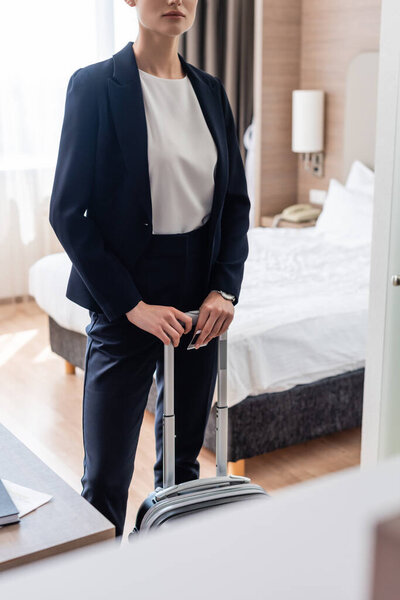 cropped view of businesswoman in suit standing with luggage in hotel room 