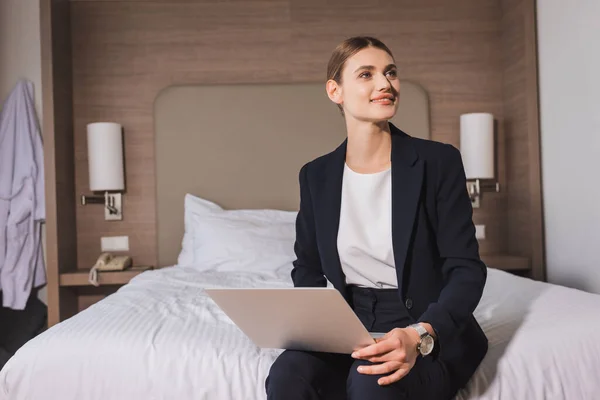スーツ姿の女性がノートパソコンを持ってベッドに座ってホテルの部屋を眺めながら — ストック写真