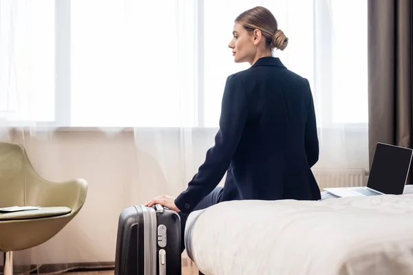 ホテルの部屋には薄型スクリーンとスーツケースを備えたラップトップの近くのベッドに座っている若いビジネスマンの女性 — ストック写真