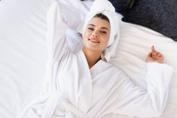 top view joyful woman in towel and white bathrobe lying on bed in hotel room