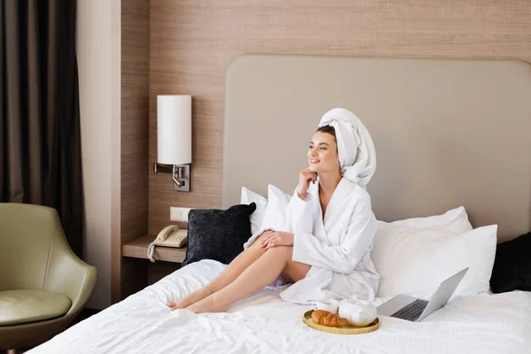 young woman in bathrobe sitting near laptop and breakfast tray on bed