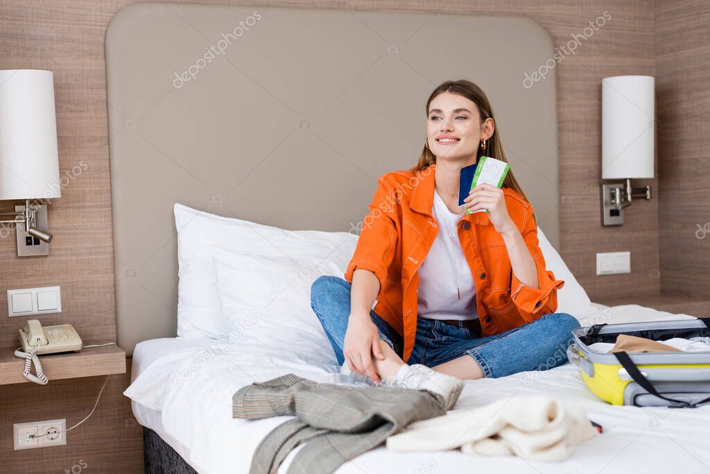 young pleased woman holding passport and air ticket near baggage and clothing on bed in hotel 