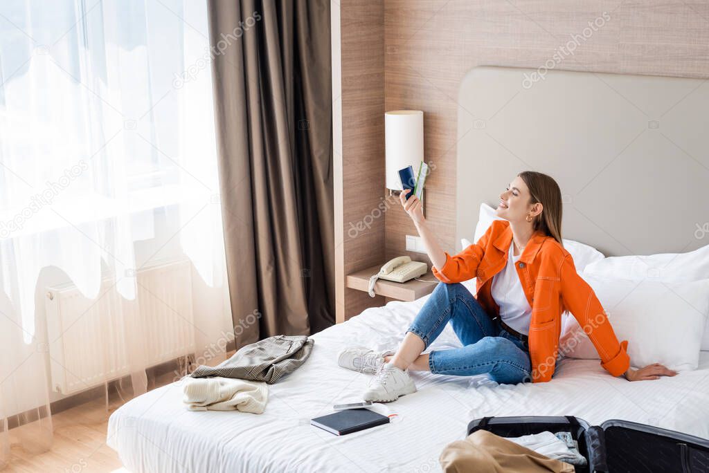 pleased woman looking at passport and air ticket near baggage and clothing on bed in hotel 