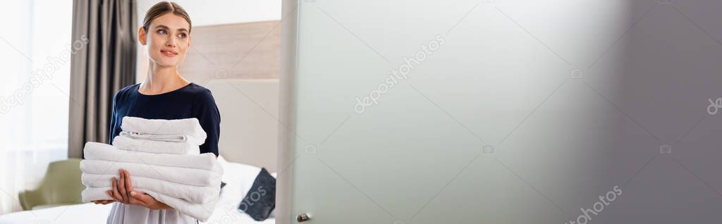 horizontal image of young maid in apron looking at camera and holding clean towels in hotel room 