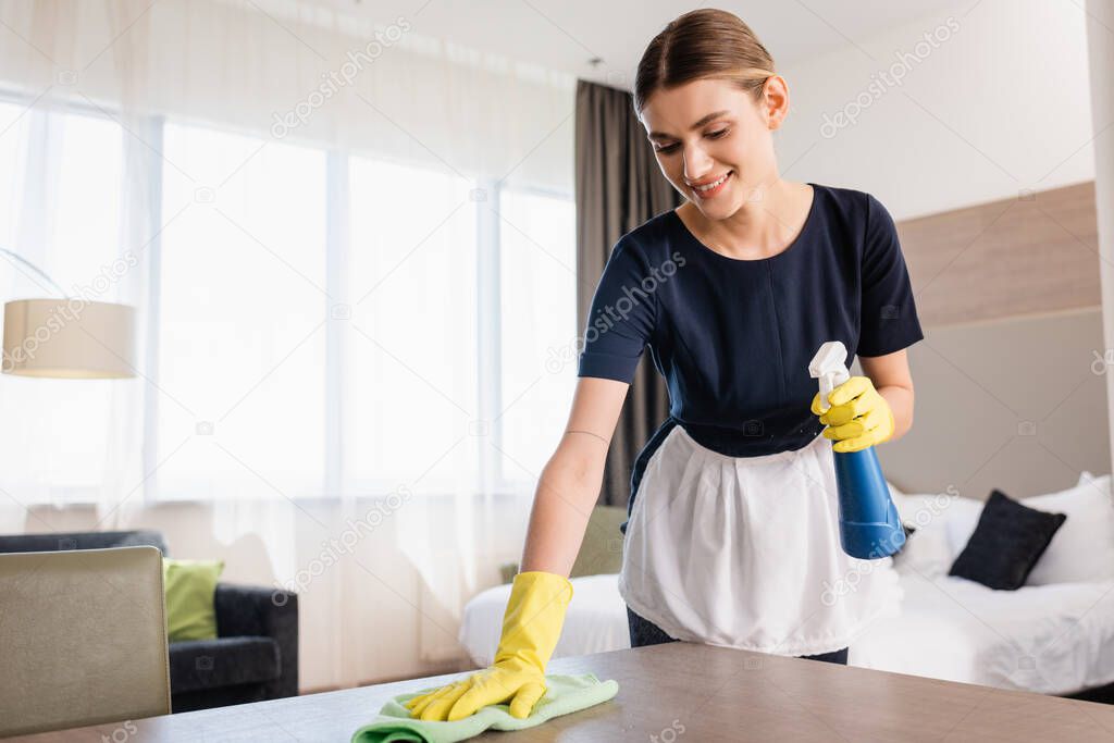 chambermaid in apron and rubber gloves holding spray bottle and rag while cleaning wooden surface in hotel room 