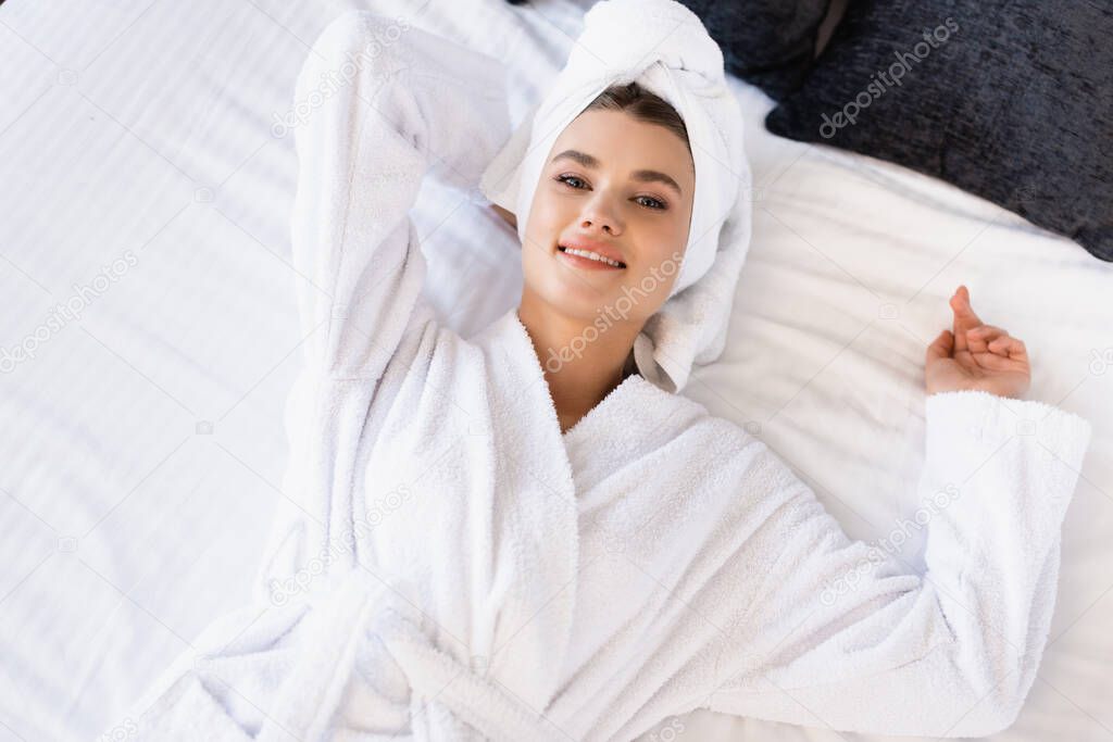 top view joyful woman in towel and white bathrobe lying on bed in hotel room