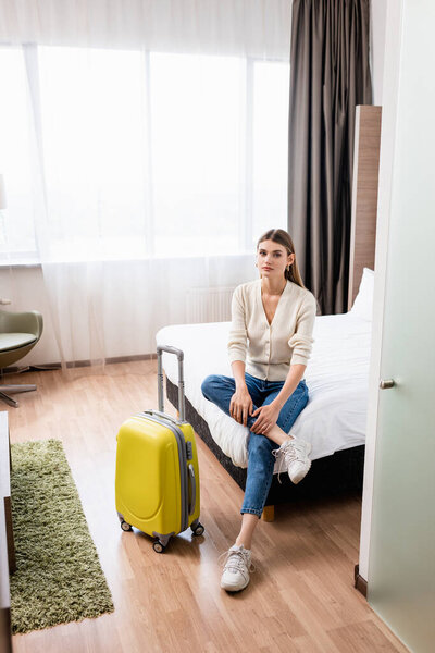 молодая женщина в джинсах сидит на кровати рядом с желтым багажом в номере отеля