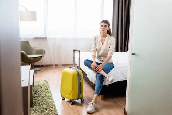 ホテルの部屋で黄色の荷物を持ってベッドの近くに座っているジーンズの若い女性の選択的な焦点 — ストック写真