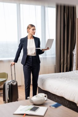 Seçici odak noktası takım elbiseli iş kadını dizüstü bilgisayarı ve bavuluyla otel odasında yatağın yanında duruyor. 