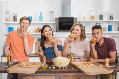 Pizza yiyen ve parti sırasında kameraya bakan çok ırklı arkadaşlar.