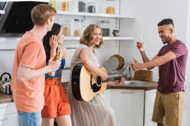 Genç bir kadın çok kültürlü arkadaşlarına gitar çalarken heyecanlı bir Afrikalı Amerikalının baş parmağını göstermesinin seçici odağı