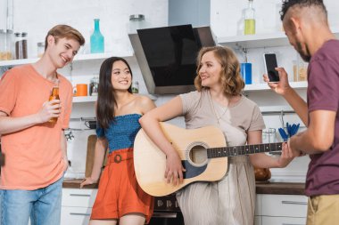Genç bir kadın çok kültürlü arkadaşlar için gitar çalarken Afro-Amerikan erkeğin akıllı telefonuyla fotoğraf çekmesinin seçici odağı.