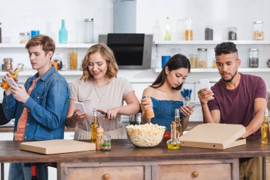 Genç çok kültürlü arkadaşlar parti sırasında bira ve patlamış mısırın yanında cep telefonlarıyla sohbet ediyorlar.