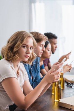 Genç bir kadın kameraya bakıyor ve çok kültürlü arkadaşlarının yanında akıllı telefondan sohbet ediyor.