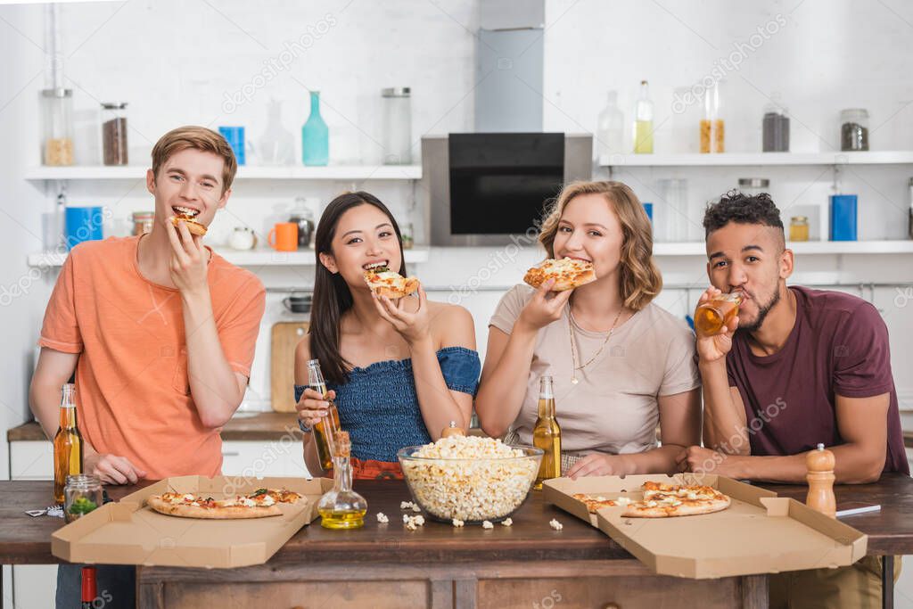 pleased multiethnic friends eating pizza and looking at camera during party