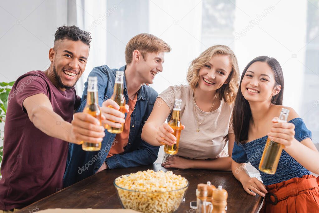 excited multicultural friends looking at camera while holding bottles of beer 