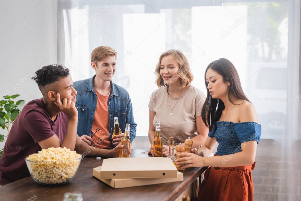 multicultural friends talking during party near beer, popcorn and pizza boxes
