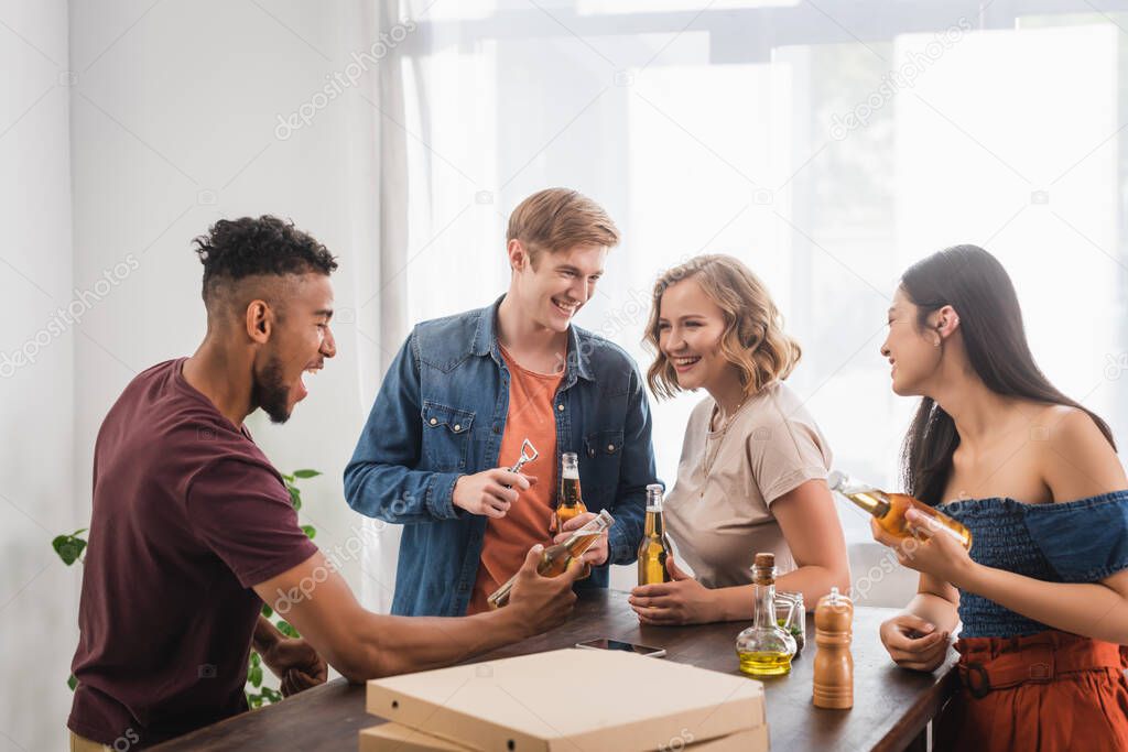 excited african american man holding bottle of beer near multiethnic friends during party