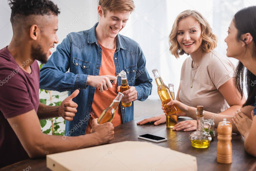 young man opening bottle of beer near multicultural friends during party