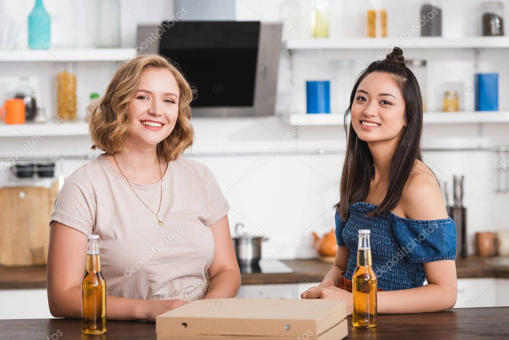 young multicultural women looking at camera near beer and pizza box on table