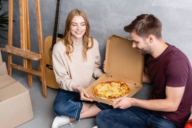 Pizza, karton kutular ve akustik gitar yanında erkek arkadaşına bakan neşeli bir kadın.