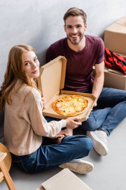 Kutuların yanında pizza yerken kameraya bakan neşeli çift.