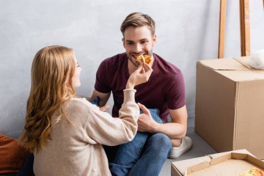 Genç kadın sakallı erkek arkadaşını kutuların yanında pizzayla besliyor.